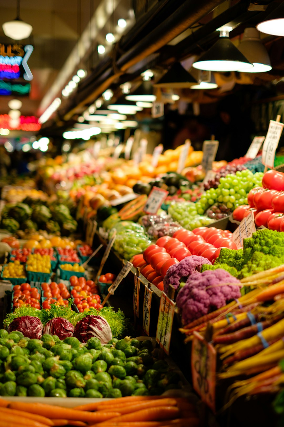 A farmers' market produce stall in late-morning light, with wooden crates of green and red cabbages, beets, radishes, hot peppers, and onions, and a hand-written chalkboard price sign.