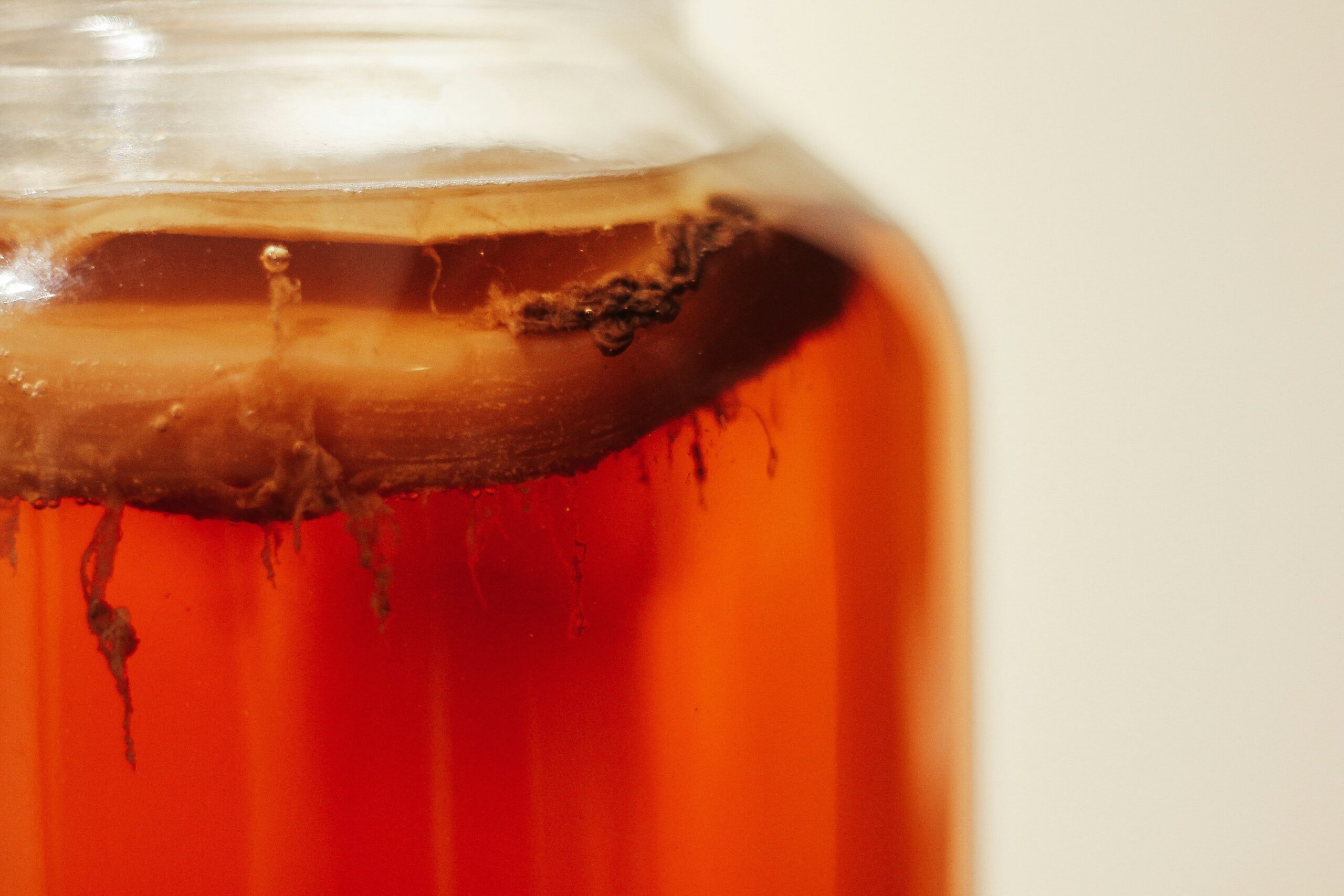 A glass jar of kombucha with a visible SCOBY floating on top, warm amber liquid, yeast strands hanging below.