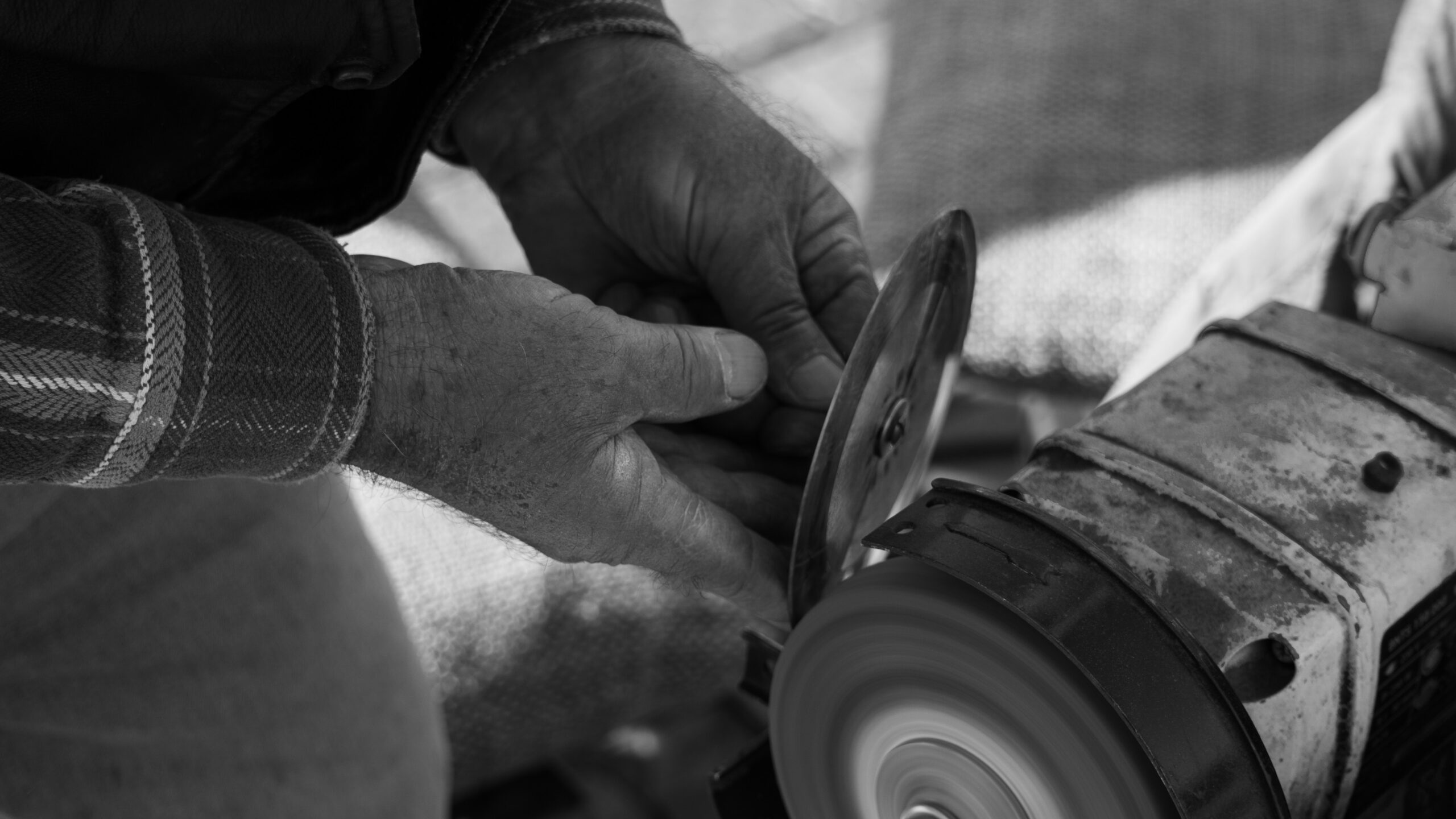 Hands guiding a tool against a bench grinder in a working shop, black and white