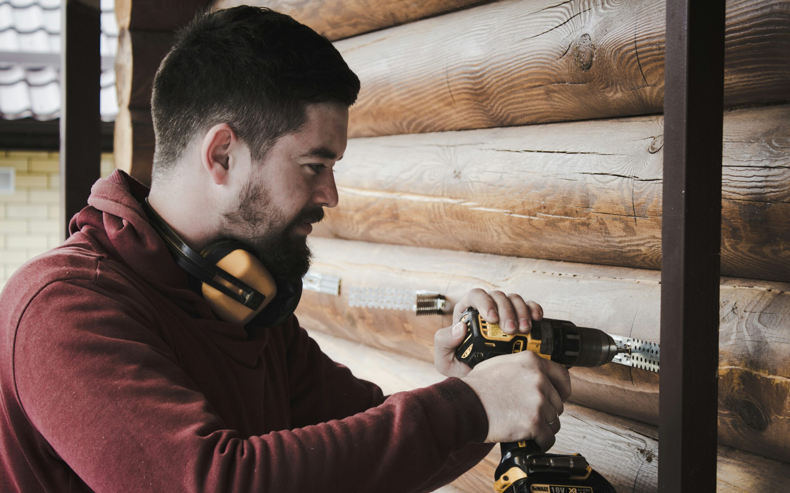 A person in work clothes running a cordless drill against a log cabin wall