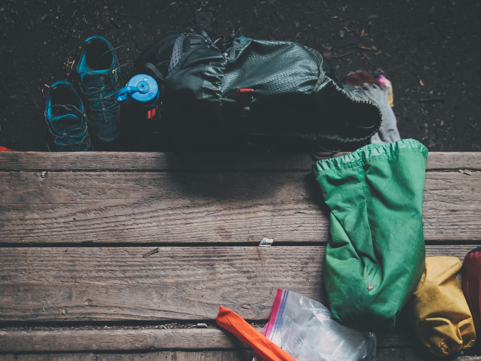 Hiking shoes, a blue water bottle, a backpacking pack, and colorful stuff sacks arranged across a forest floor and weathered wooden planks