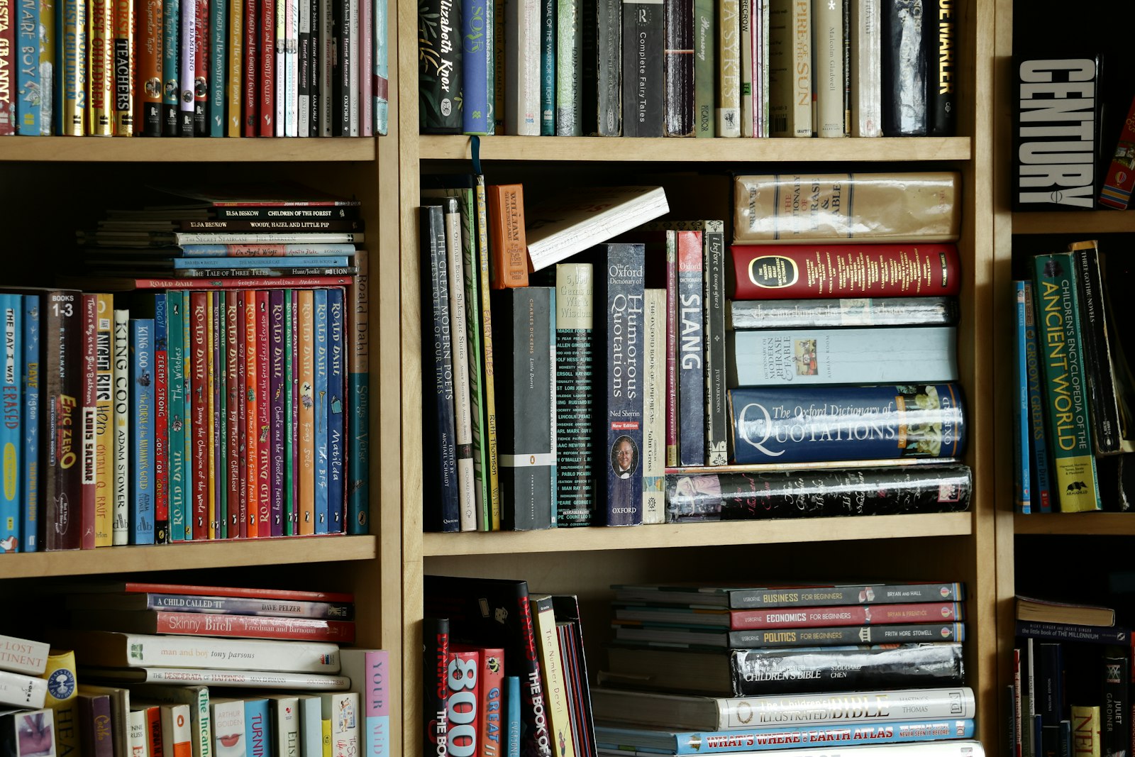 A light wooden home bookshelf packed tightly with physical books of many sizes and colors, arranged vertically and in stacks, in soft warm light