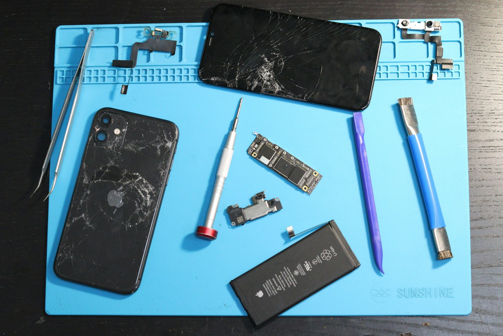 A disassembled smartphone with its screen, battery, and circuit board laid out on a blue silicone repair mat with precision screwdrivers and tweezers