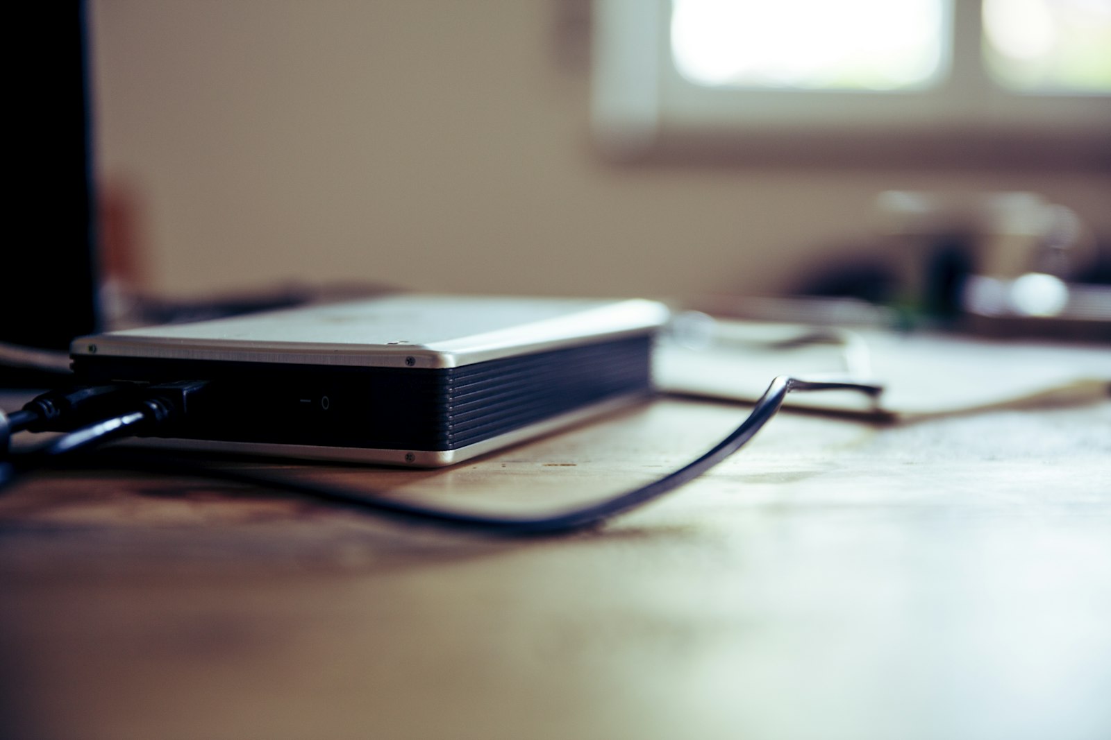 A silver and black external hard drive enclosure with power and data cables connected, resting on a wooden desk in soft window light