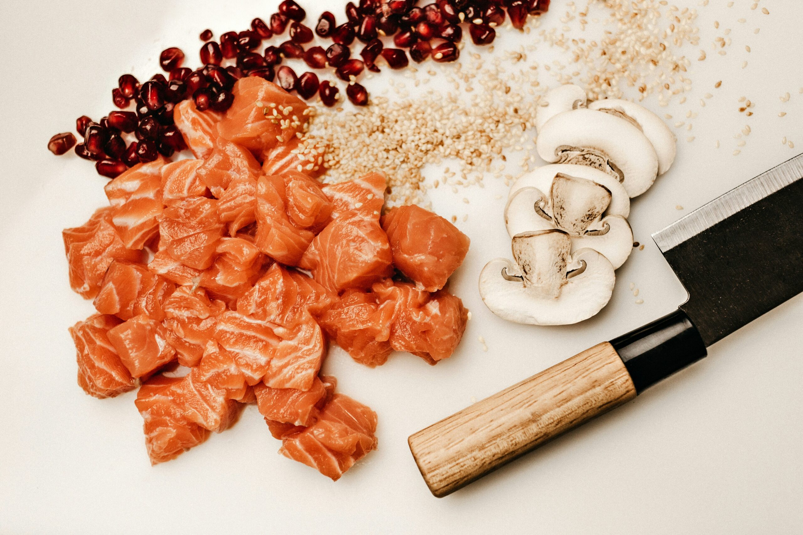 Diced raw salmon, sliced mushrooms, pomegranate seeds, and sesame arranged on a white surface
