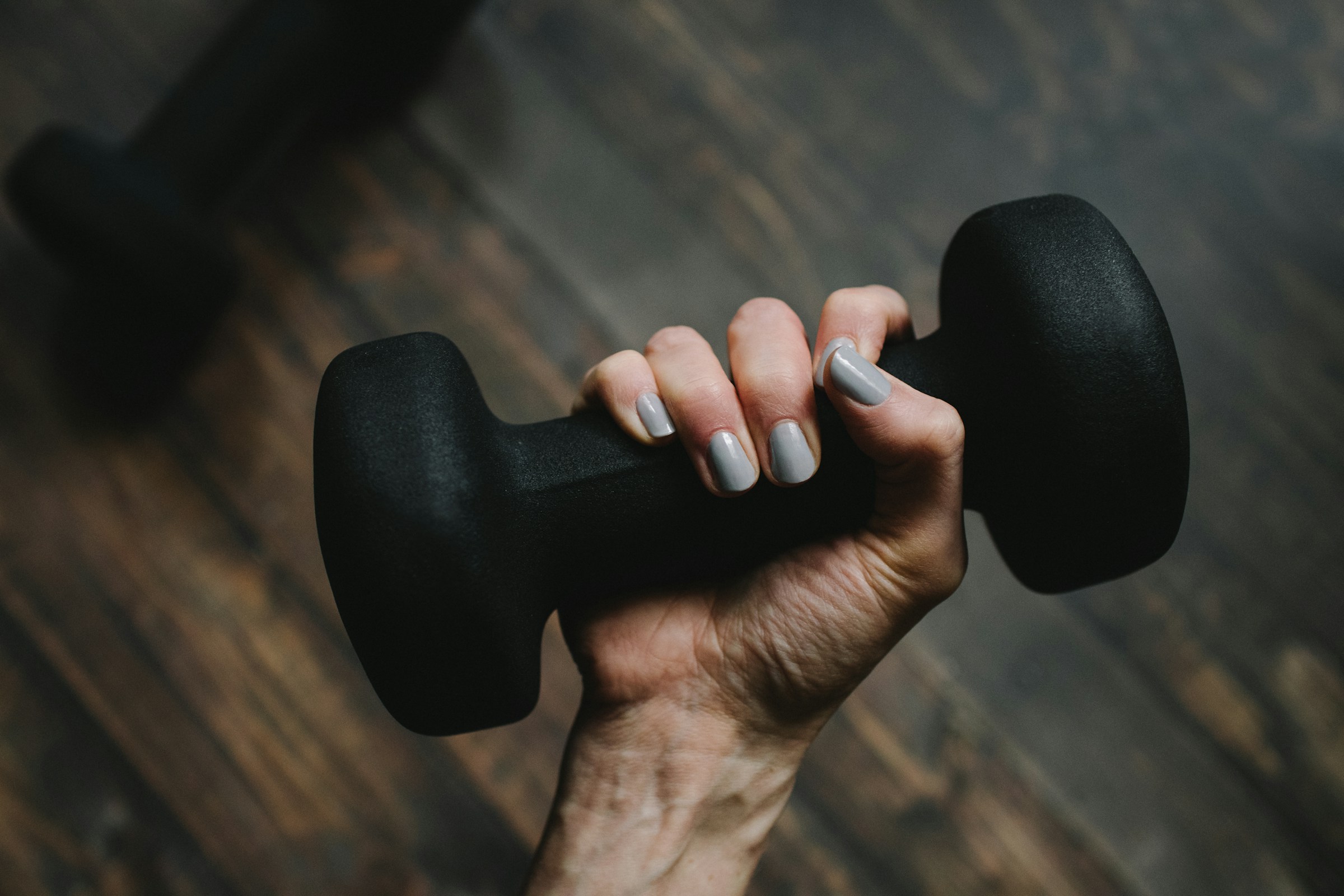 A hand gripping a small neoprene dumbbell on a wooden floor at home